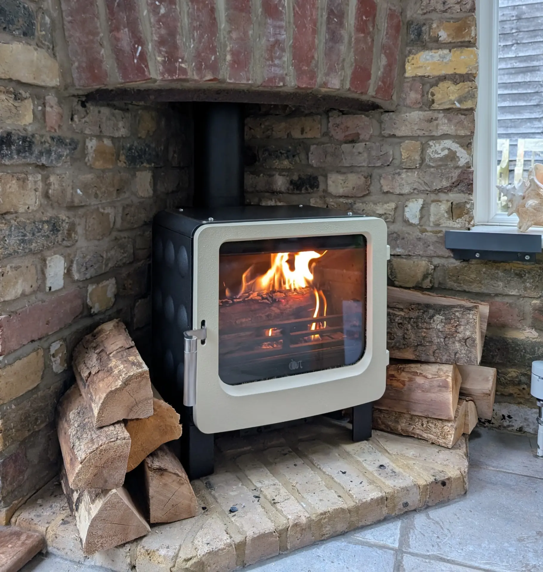 Wood burning stove installation in Windsor, cream-fronted stove alight in a period brick inglenook with stacked logs